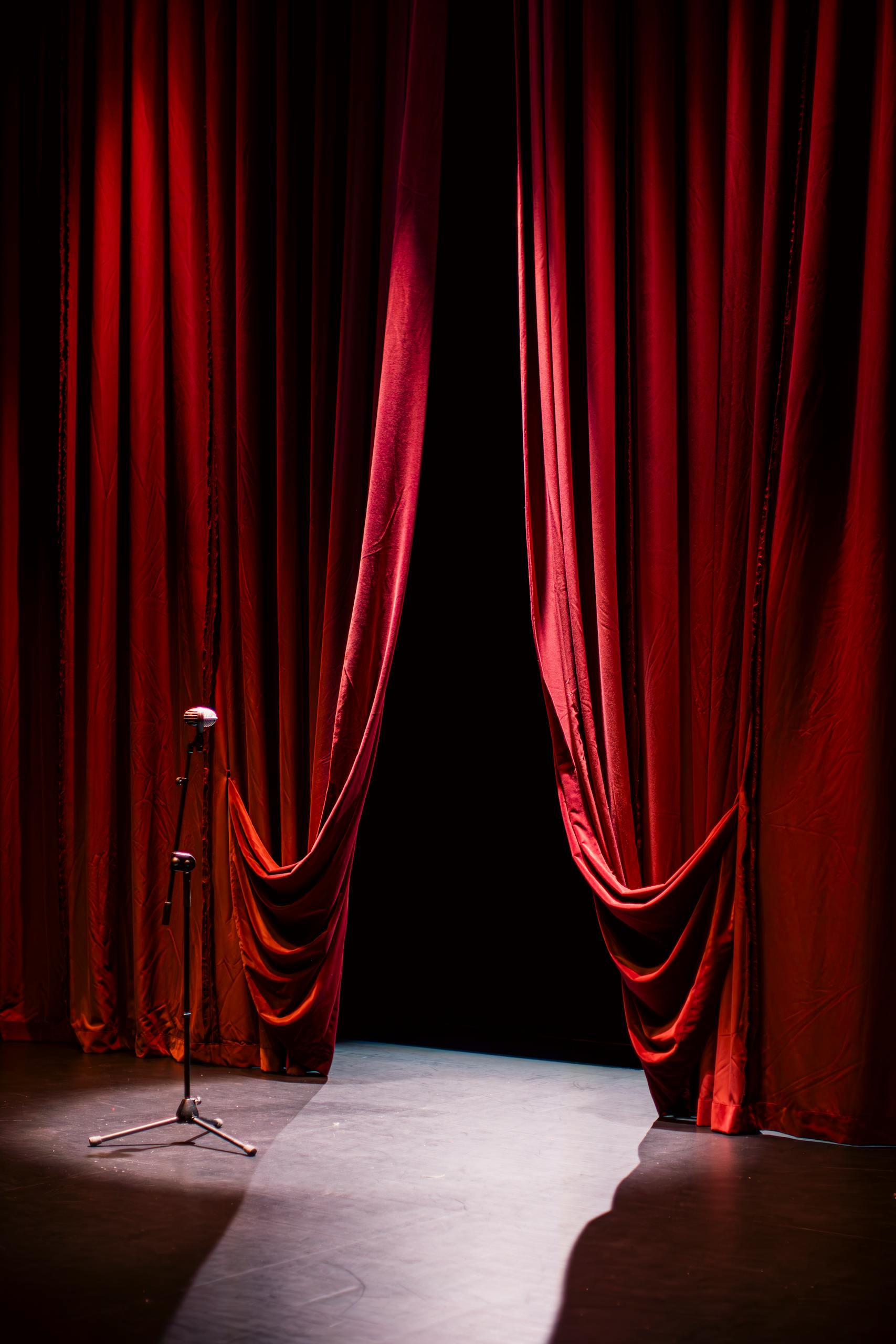 A theater stage with closed red curtains and a single microphone in the spotlight, creating a dramatic ambiance.